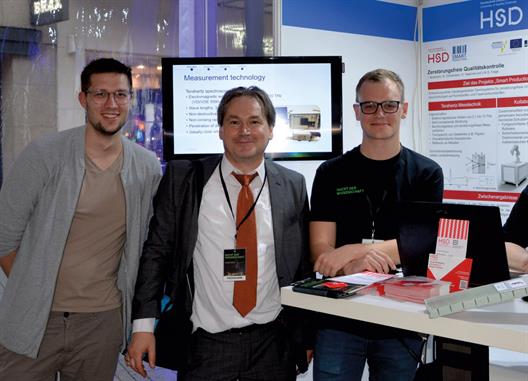 Die Arbeitsgruppe von Prof. Dr.-Ing. Volker Feige (Mitte) und Prof. Dr. Hartmut Haehnel vom Fachbereich Elektro- und Informationstechnik stelle das Interreg-Projekt „Smart Production“ vor, das die Qualität von Beschichtungen mit einem automatisierten robotergestützten System ermöglichen soll. Das Team erläuterte am Informationsstand die Algorithmen für die zerstörungsfreie Untersuchung von Schutzbeschichtungen mittels THz-Messtechnik, welche insbesondere für Beschichtungssysteme auf Kunststoffen und Verbundstoffen erforscht werden. Das große Interesse der Besucher*innen an der Veranstaltung zeigte sich durch zahlreiche Gespräche, die bis Mitternacht stattfanden. 